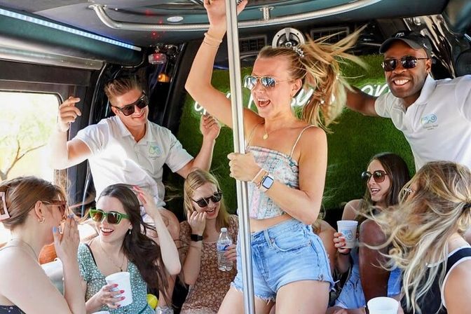 Group of young adults partying inside a daytime party bus, woman dancing on a pole in denim shorts as friends laugh, sip drinks and wear sunglasses — energetic summer celebration