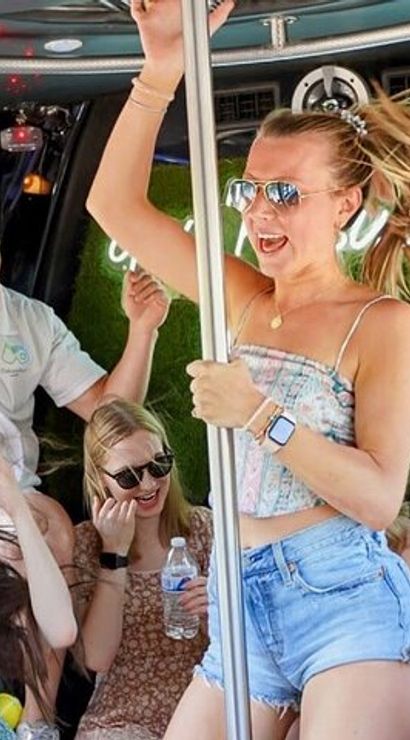 Group of young adults partying inside a daytime party bus, woman dancing on a pole in denim shorts as friends laugh, sip drinks and wear sunglasses — energetic summer celebration