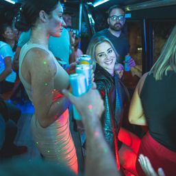 Neon-lit party bus at night with friends, a smiling woman reaching for a canned drink while others hold beverages and colorful disco lights sparkle.