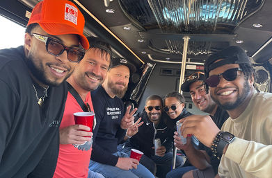 Seven friends smiling inside a party bus limo, wearing sunglasses and caps, holding red cups and canned drinks around a chrome pole during a daytime celebration.