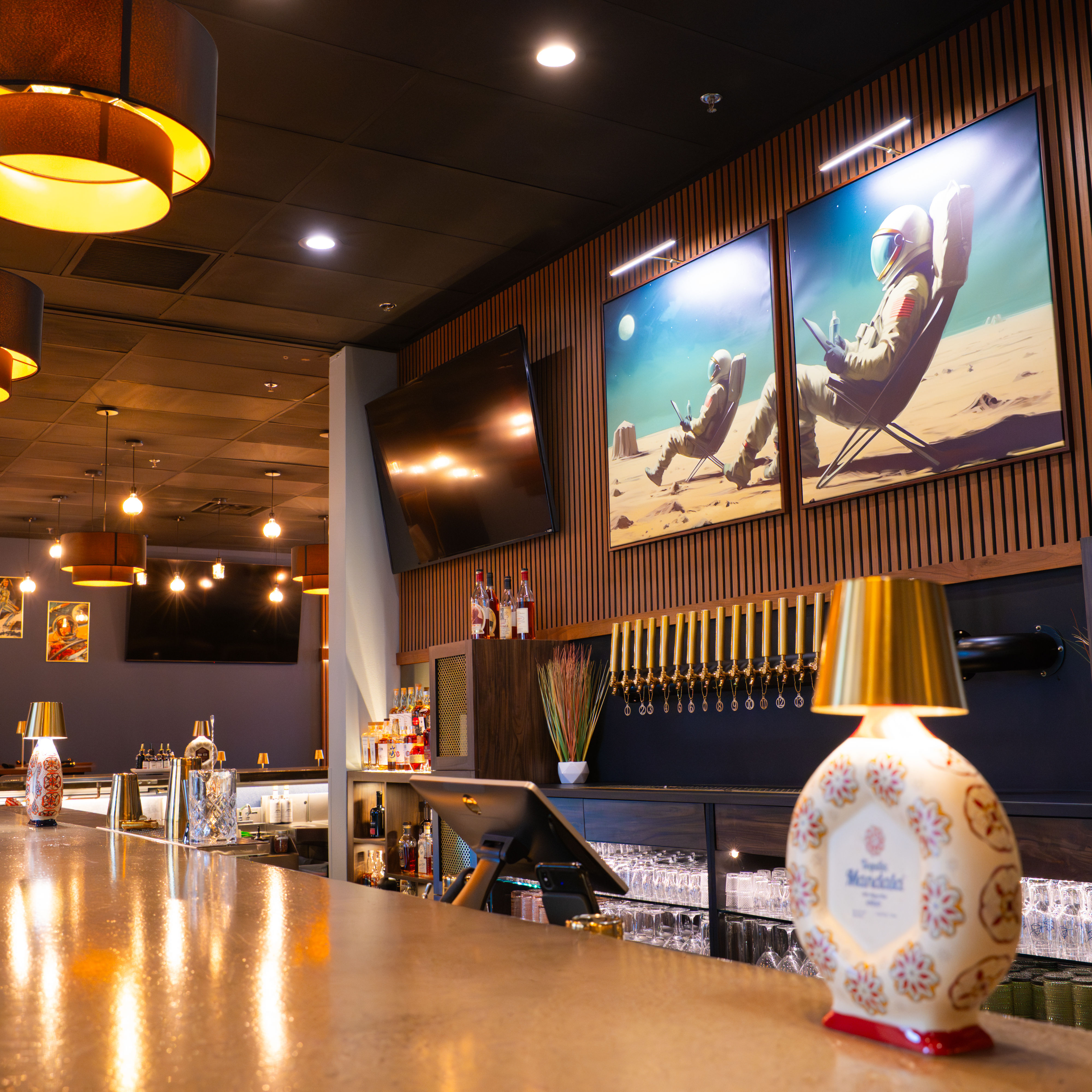 Modern upscale bar interior with long polished counter, brass beer taps, glassware and bottles, warm pendant lights and space-themed astronaut paintings