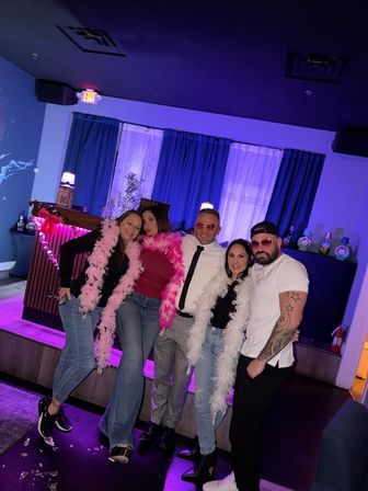 Five adults posing in a purple-lit lounge bar in front of a curtained DJ booth, wearing feather boas and casual outfits — playful nightlife group photo.