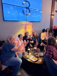 Group of friends toasting with shot glasses around a low table of small plates and cocktails in a cozy indoor cocktail lounge, under a large blue wall screen showing a number 3 and a pink feathered lamp.