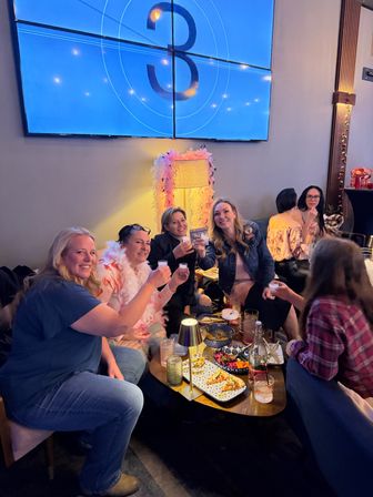 Group of friends toasting with shot glasses around a low table of small plates and cocktails in a cozy indoor cocktail lounge, under a large blue wall screen showing a number 3 and a pink feathered lamp.