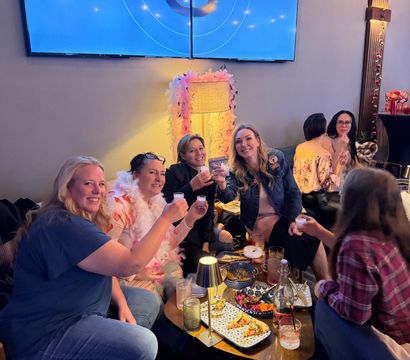 Group of friends toasting with shot glasses around a low table of small plates and cocktails in a cozy indoor cocktail lounge, under a large blue wall screen showing a number 3 and a pink feathered lamp.