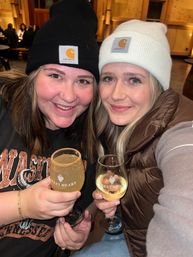 Two friends in knit beanies smile in a cozy wood-paneled winery tasting room, each holding a stemmed glass of white wine during a wine tasting.