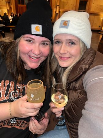 Two friends in knit beanies smile in a cozy wood-paneled winery tasting room, each holding a stemmed glass of white wine during a wine tasting.