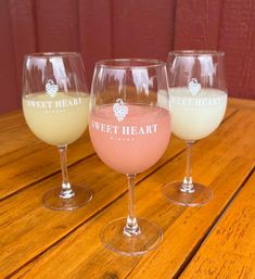 Three stemmed wine glasses with pink and pale yellow frozen wine slushies on a wooden patio table in front of red siding.
