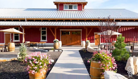 Sunlit red barn-style building with metal roof and double wooden doors, paved walkway flanked by wooden barrel planters of pink flowers, outdoor picnic tables with umbrellas and landscaped rock beds creating a rustic patio entrance.