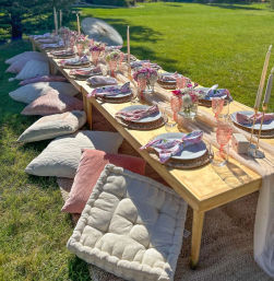 Boho outdoor garden picnic on a sunny lawn: low wooden table set with woven chargers, pink glassware, floral centerpieces, taper candles and pastel floor cushions.