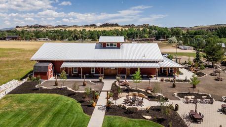 Aerial view of a red barn-style event venue with a metal roof, landscaped patios, picnic tables and a circular firepit area, set in sunny rural countryside with grassy fields and distant rocky hills