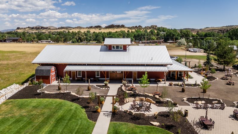 Aerial view of a red barn-style event venue with a metal roof, landscaped patios, picnic tables and a circular firepit area, set in sunny rural countryside with grassy fields and distant rocky hills