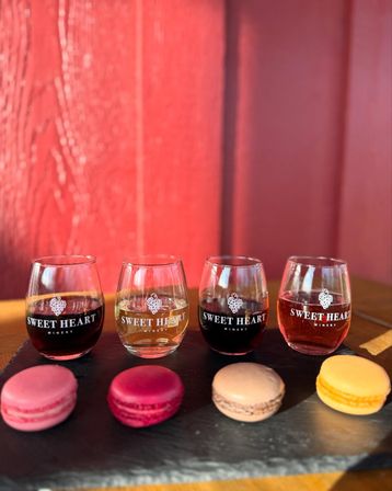 Wine tasting flight: four stemless glasses of red, white and rosé on a slate board paired with four colorful macarons against a sunlit red wooden wall.