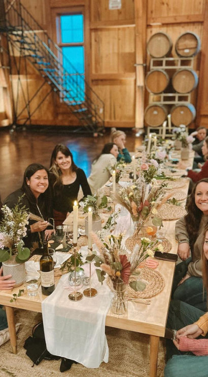 Group of women seated at a low wooden table in a rustic barrel-room tasting space, enjoying a cozy candlelit dinner with wine bottles, dried floral centerpieces and woven placemats