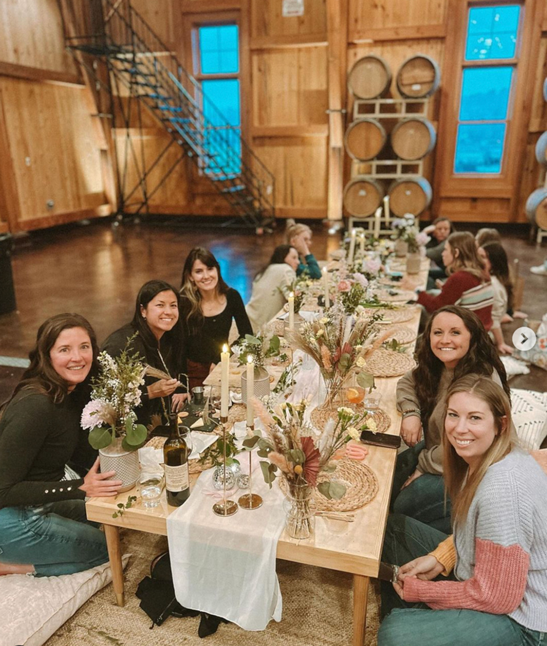 Group of women seated at a low wooden table in a rustic barrel-room tasting space, enjoying a cozy candlelit dinner with wine bottles, dried floral centerpieces and woven placemats