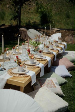 Sunlit boho garden picnic with a long low wooden table set on grass—woven chargers, white plates, small floral potted arrangements, taper candles, crystal glassware, and pastel floor cushions for seating.