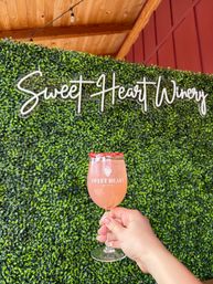 Hand holding a pink rosé cocktail in a stemmed wine glass with a red fruit rim, posed against a lush green hedge photo wall and neon script sign at an outdoor winery patio.