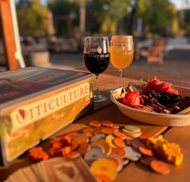 Sunlit winery patio scene with two stemmed glasses (red wine and amber brew) on a wooden table beside a charcuterie platter, scattered board game tokens and a vineyard-themed board game box.