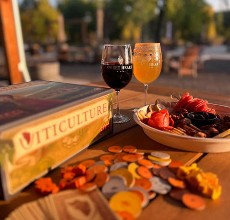 Sunlit winery patio scene with two stemmed glasses (red wine and amber brew) on a wooden table beside a charcuterie platter, scattered board game tokens and a vineyard-themed board game box.