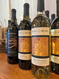 Close-up of assorted wine bottles on a wooden counter, spotlighting a 2021 Tempranillo and a White Wine Blend from Columbia Valley, Washington with warm orange-gold label artwork.