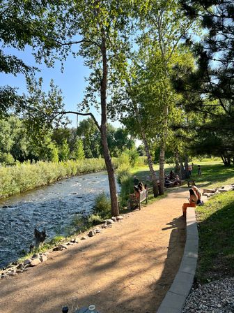 Sunny riverside park with a winding dirt trail, tall green trees, benches and people relaxing along a shallow rocky creek on a summer afternoon