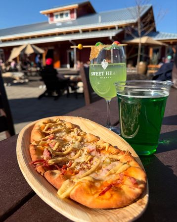 Sunny outdoor patio at a rustic tasting room featuring a flatbread on a wooden platter and two vibrant green drinks—one in a stemmed glass with candy garnish—on a picnic table.