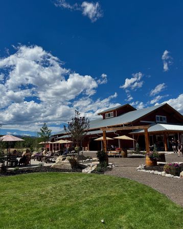 Sunny outdoor patio at a rustic barn-style venue with wooden beams, umbrellas, barrel planters and seating on a green lawn under a vibrant blue sky with fluffy clouds and distant mountain views.