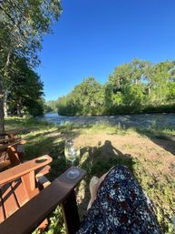 Riverside park view from a chair — wine glass on the armrest, floral-skirted legs, grassy riverbank, trees and bright blue sky