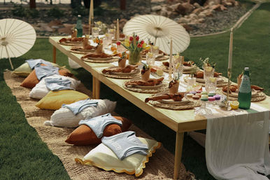 Whimsical boho garden picnic: low wooden table on grass with woven placemats, knotted linen napkins, crystal glasses, taper candles, tulip centerpiece, colorful macarons, sparkling water, paper parasols and floor cushions for casual outdoor seating.