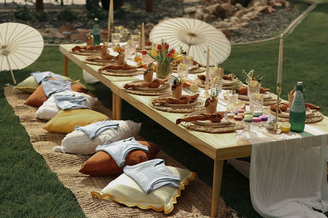 Whimsical boho garden picnic: low wooden table on grass with woven placemats, knotted linen napkins, crystal glasses, taper candles, tulip centerpiece, colorful macarons, sparkling water, paper parasols and floor cushions for casual outdoor seating.