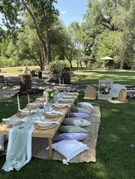 Boho-chic outdoor picnic on a green lawn under shaded trees — low wooden table set with woven chargers, cloth napkins, pastel glassware, taper candles and a floral centerpiece, surrounded by pastel cushions on a woven rug with park-style picnic tables and umbrellas in the background.