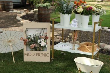 Outdoor garden picnic decor on grass: white paper parasol, framed floral arrangement, two-tier metal cart styled as a flower bar with white buckets of mixed blooms, wicker picnic basket and small decorative tub.