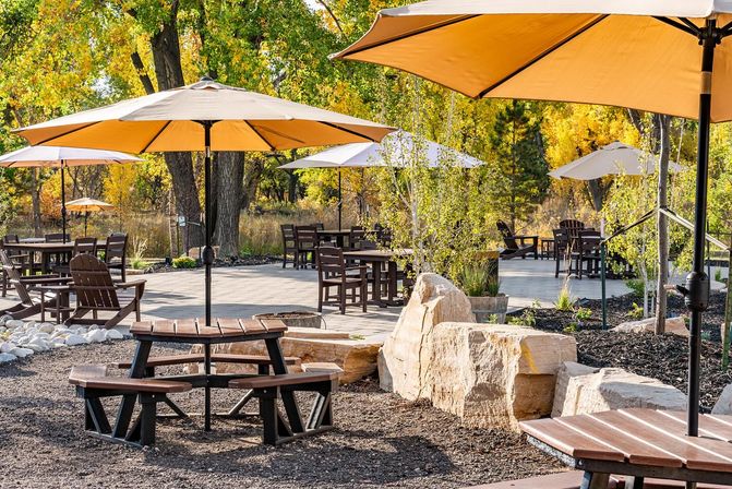 Sunlit outdoor patio with wooden picnic tables, circular bench seating and beige umbrellas on a stone-lined deck surrounded by golden autumn trees and rock landscaping
