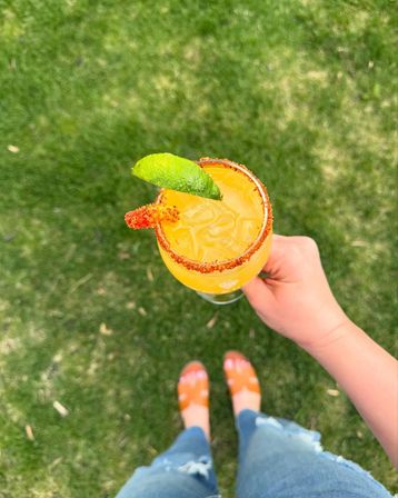 Hand holding an icy bright orange citrus cocktail with a chili-salt rim and lime wedge, overhead shot over green backyard grass with casual sandals and ripped jeans visible.