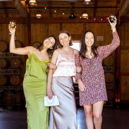 Three women in colorful dresses toasting with wine glasses inside a wooden winery barrel room, smiling in front of stacked oak barrels and string lights.