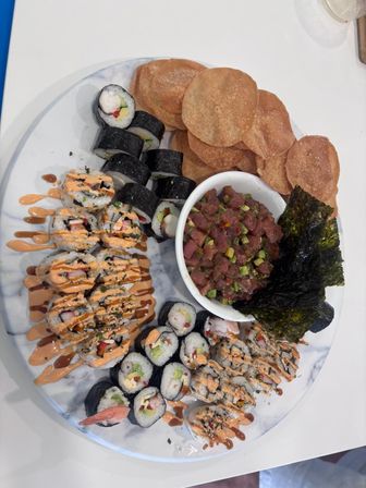 Overhead view of an Asian-fusion sushi platter on a white marble plate featuring assorted maki rolls drizzled with spicy mayo and eel sauce, a bowl of diced tuna poke with avocado, roasted seaweed sheets, and crispy wonton chips.