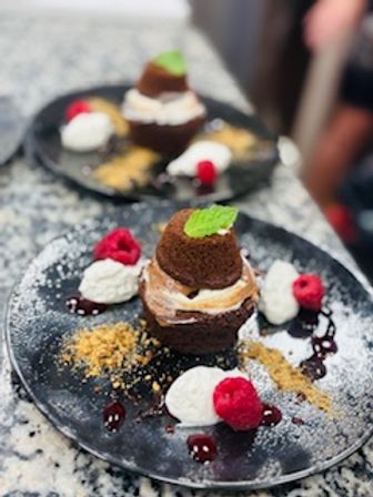 Mini chocolate lava cake topped with a mint leaf, plated with whipped cream dollops, fresh raspberries, crumbled biscuit, chocolate sauce and powdered sugar on a black plate over a granite countertop — elegant restaurant dessert