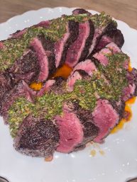 Close-up of sliced medium-rare seared steak drizzled with green chimichurri sauce and juices on a white plate