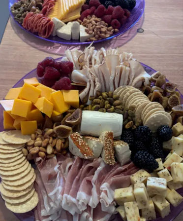 Gourmet charcuterie platters on purple plates: cubed cheddar and herbed cheeses, a goat cheese log, thinly sliced cured meats, round crackers, dried figs, pistachios and peanuts, sesame-coated breadsticks, fresh blackberries and raspberries on a wooden table.