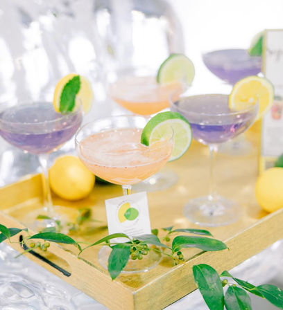 Gold serving tray with pastel peach and purple coupe cocktails, each garnished with lime or lemon slices, fresh lemons and green leaves for a bright citrus party display