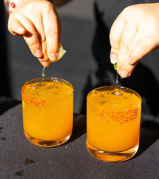 Two bright orange citrus cocktails on ice with chili-salt rims as hands squeeze fresh lime over them