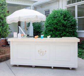 White mobile outdoor bar on casters with a fringed umbrella and colorful cocktails on a suburban front-porch patio framed by green shrubs