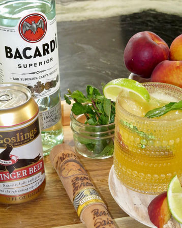 Peach‑colored rum cocktail with lime wedge and mint on a wooden board, surrounded by a white rum bottle, ginger beer can, a cigar, and fresh peaches.