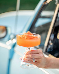 Hand holding a ribbed coupe glass with a bright orange cocktail topped with a peach flower garnish, blurred pale blue car in the background.