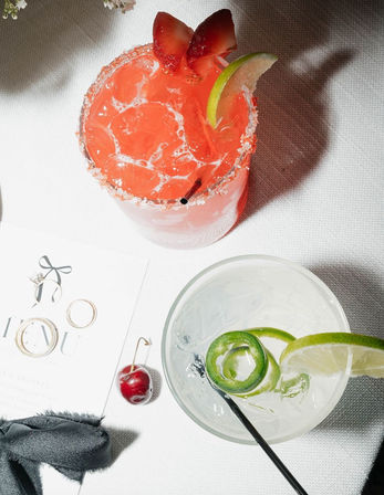 Top-down view of two craft cocktails on a white tablecloth: a salt-rimmed pink strawberry-lime drink with ice and strawberry garnish, and a clear jalapeño-lime cocktail with ice, lime wheel and straw, with a cherry and gold rings nearby.