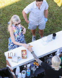 Overhead view of a bartender handing a tropical cocktail to a woman in a floral dress and lei at a pop-up outdoor bar on a grassy backyard during a summer luau.