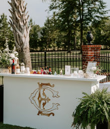 Outdoor white wedding bar with gold monogram on a lawn, stocked bottles and mixers, floral arrangements and ferns, set by a palm tree, brick pillar and wrought-iron fence in a garden