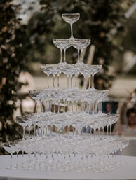 Pyramid champagne tower of stacked clear coupe glasses on a table at an outdoor wedding reception