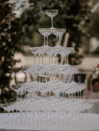 Pyramid champagne tower of stacked clear coupe glasses on a table at an outdoor wedding reception