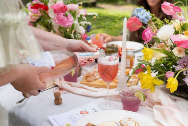 Hand pouring sparkling rosé into a frothy pink cocktail on a flower-filled outdoor brunch table with colorful spring blooms and appetizers.